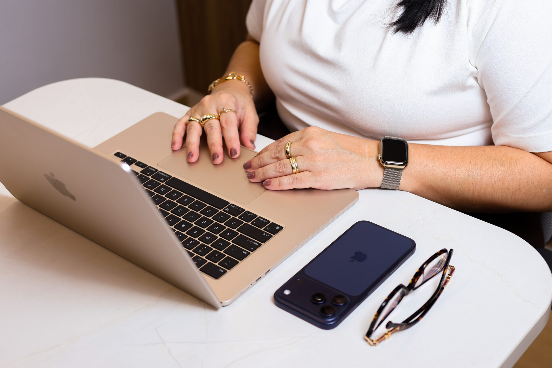 Mãos de Patrícia Bastos sobre o notebook, com celular e óculos sobre a mesa