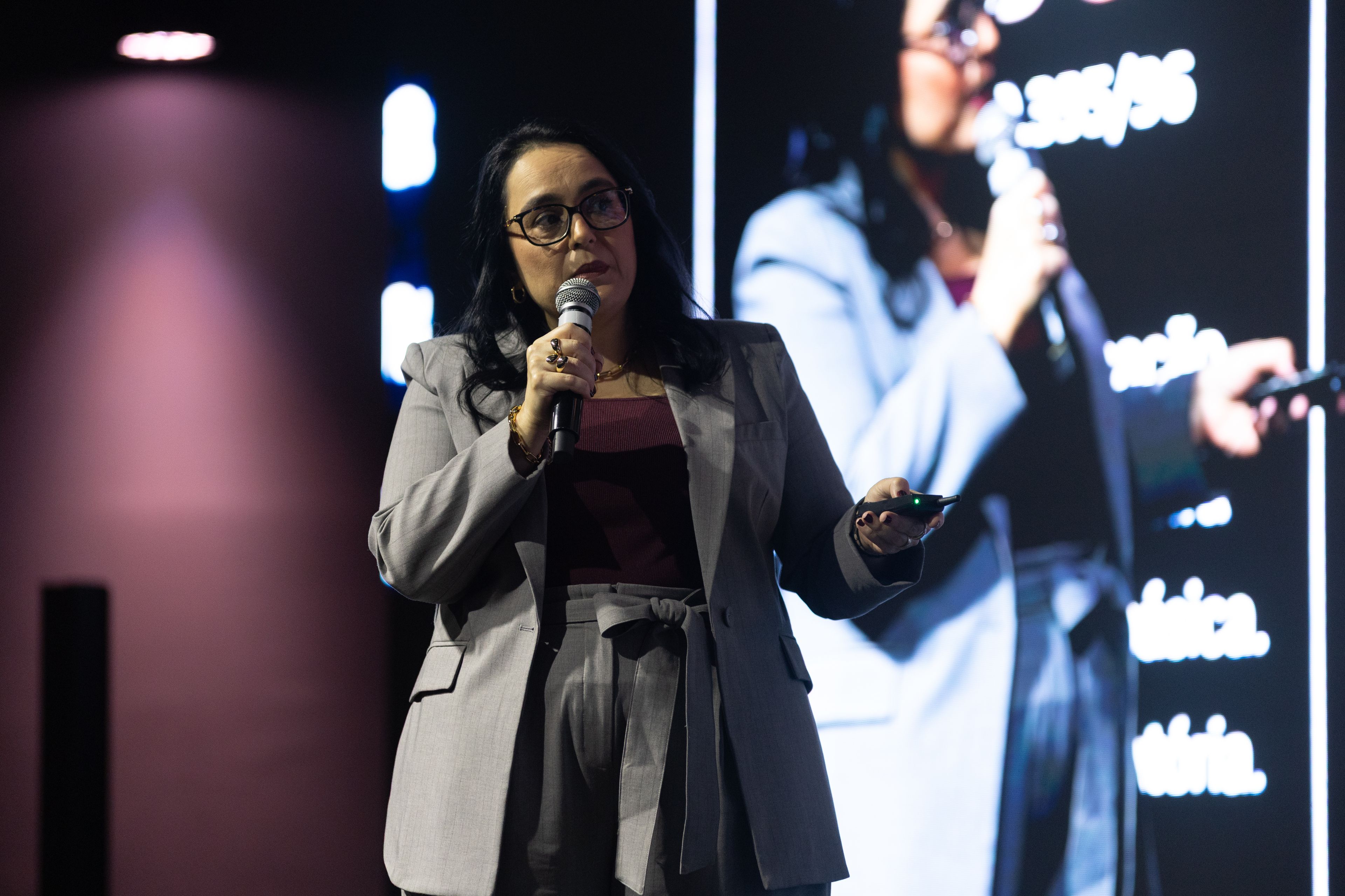 Patrícia Bastos palestrando no palco, com microfone e apresentação ao fundo